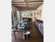 Ein moderner Büroraum mit dunklem Holzboden, einer langen Glasarbeitsfläche mit mehreren Bürostühlen und Schreibtischlampen. An den Wänden hängen Kunstwerke, und Regale mit Büromaterialien säumen den rechten Bereich; die Decke zeigt freiliegende Balken.