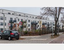 Mehrfamilienhaus in moderner Bauweise mit weißer Fassade und dunklen Balkonen, umgeben von einem Zaun und einer kleinen bepflanzten Gartenanlage. Im Vordergrund sind ein geparktes Auto, Straßenschilder, ein Gehweg und eine Baumreihe sichtbar.