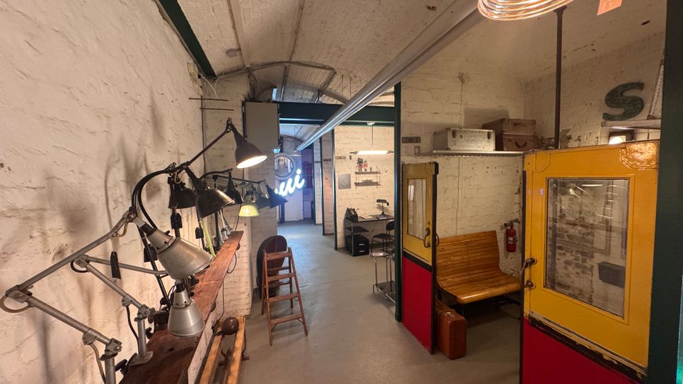 Ein industriell gestalteter Innenraum mit weiß gestrichenen Backsteinwänden, gewölbter Decke und sichtbaren Lampen, die an einer Holzleiste montiert sind. Rechts steht eine gelb-rot gestrichene alte Schienenfahrzeugkabine mit Holzbänken, während der Raum im Hintergrund einen Arbeitsplatz und weitere Gegenstände zeigt.