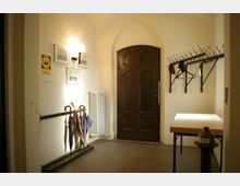 Eingangsbereich mit gewölbter Decke, einer dunkelbraunen Holztür, einem Schirmständer mit Schirmen, einer Garderobe mit Haken und einem Heizkörper an der Wand. Rechts neben der Tür steht ein kleiner Tisch und ein Behälter mit der Aufschrift "Kirche".