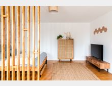 Schlafzimmer mit minimalistischem Design; ein Bett mit Holzrahmen und blauer Bettwäsche, eine dekorative Holzgitter-Trennwand, ein Sideboard aus Holz mit TV, ein Schrank mit Rattan-Fronten und Pflanzen sowie ein heller Teppich auf einem Holzboden.