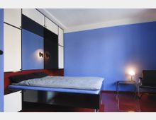 Schlafzimmer mit blauer Wand und roten Böden. Ein Bett mit schwarzem Rahmen, darüber schwarz-weiße Hängeschränke, daneben ein kleiner Beistelltisch mit Lampe und ein schwarzer Stuhl.