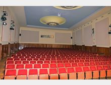 Ein Theater- oder Kinosaal mit Reihen roter Sitzplätze, holzverkleideten Wänden und einer hellen Decke mit zwei großen, dekorativen Lampen. An den Seiten sind Lautsprecher und Beleuchtung installiert.