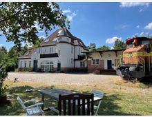 Weißes Gebäude mit rotem Ziegelsockel und dunklem Schrägdach, daneben eine stillgelegte, rostige Lokomotive. Im Vordergrund stehen Gartenmöbel auf einer Wiese, umgeben von Bäumen, unter blauem Himmel mit einigen Wolken.