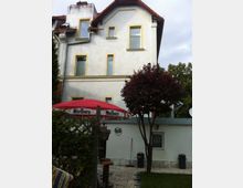 Zweistöckiges Wohnhaus mit weißer Fassade und gelben Fensterrahmen, einem roten Satteldach und mehreren Fenstern. Im Vordergrund befindet sich ein Gartenbereich mit einem roten Sonnenschirm, Stühlen, einem Baum und einer kleinen Terrasse aus Pflastersteinen.