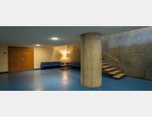 Ein moderner, minimalistischer Innenraum mit Betonwänden, blauem Bodenbelag und einem zentralen Rundpfeiler. Ein Bereich mit blauen Sofas und einem Beistelltisch steht links, während eine freitragende Treppe mit Holzgeländer rechts sichtbar ist; eine Holztür führt zu einem Konferenzraum.
