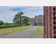 Backsteinbau mit großen Fenstern im klassischen Stil, umgeben von einer gepflegten Grünfläche mit Bäumen und Hecken; im Vordergrund eine asphaltierte Wegführung und Laternen. Links am Horizont sind Felder und ein weiteres Bauwerk zu sehen.