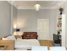 Wohnzimmer mit heller Wandfarbe und hohen Decken, verziert mit Stuckelementen. Eine braune Ledercouch, ein Sofa mit Bettlaken, ein Bücherregal und ein kleiner Fernseher auf einem niedrigen Schrank prägen den Raum; eine Pendelleuchte hängt zentral von der Decke.