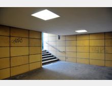 Ein unterirdischer Gang mit gelb gefliesten Wänden, vereinzelt mit Graffiti versehen. Eine Treppe mit Metallgeländer führt nach oben zu einer helleren Außenöffnung, und rechteckige Deckenleuchten beleuchten den Raum.