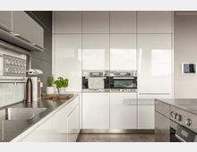 Moderne Küche mit hellen, glatten Hochglanzfronten und integriertem Backofen. Ein Edelstahlspülbecken und Arbeitsflächen erstrecken sich entlang der linken Seite, dekoriert mit Kräutertöpfen und Zubehör, während die rechte Seite einen Herd mit Bedienknöpfen zeigt.