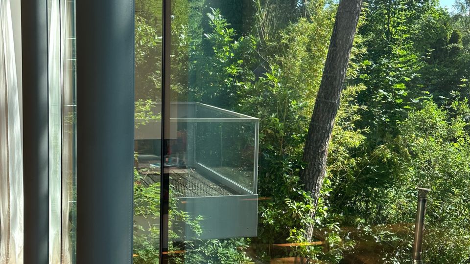 Blick durch eine bodentiefe Glasfront eines modernen Hauses auf eine Terrasse mit Glasgeländer und Holzdeck, umgeben von dichtem Grün. Im Garten sind hohe Bäume, Sträucher, Steinplatten sowie eine kleine Feuerstelle und ein rundes Becken mit Metallrohr zu sehen.