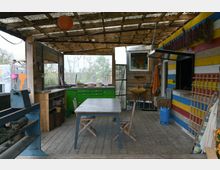 Überdachter Außenbereich mit Holzboden und Holzwänden. Im Raum befinden sich ein Tisch mit zwei Stühlen, eine grüne Küchenzeile, eine gelbe Spüle und bunte Wandverkleidungen mit blau-gelb-roten Streifen.