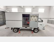 Ein Barkas B1000 Kleintransporter in einer hellen Garage mit offenem Seitentürbereich, der den Innenraum mit roten Sitzpolstern zeigt. Das Fahrzeug steht auf roten Unterstellböcken, und die Garage hat weiße Wände und helle Deckenleuchten.