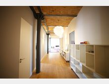 Langer, heller Flur mit Holzboden, weißen Wänden und einer Decke aus unverputzten Ziegeln, verstärkt durch schwarze Stahlträger. Rechts befindet sich ein weißes Regal, und eine moderne Hängelampe ist zentral positioniert.