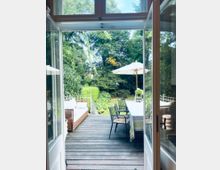 Blick durch geöffnete Terrassentüren auf eine Holzterrasse mit einem langen Tisch, Stühlen und einem Sonnenschirm; im Hintergrund ein Garten mit dichter Vegetation und Büschen.