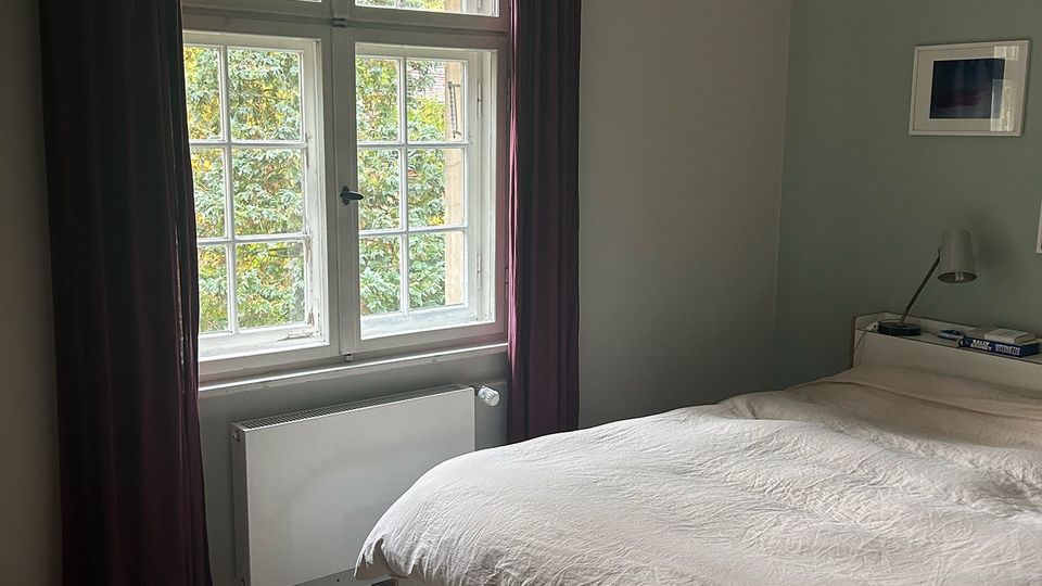 Helles Schlafzimmer mit großem Sprossenfenster und dunklen Vorhängen, darunter ein Heizkörper; draußen sind Bäume zu sehen. Ein weißes Bett mit Bettdecke steht neben einem kleinen Nachttisch, an der Wand hängen gerahmte Bilder und eine geflochtene Deckenlampe.