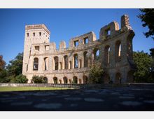 Ruine eines steinernen Gebäudes mit Rundbögen und einem hohen Turm, umgeben von grünem Rasen und Bäumen. Der blaue Himmel bildet den Hintergrund.