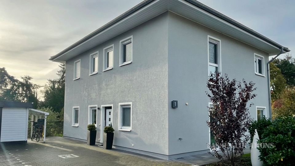 Zweistöckiges, modernes Wohnhaus mit hellgrauer Fassade und flachem Walmdach, mit mehreren rechteckigen Fenstern und einem Eingangsbereich mit Topfpflanzen. Davor liegt eine gepflasterte Einfahrt; links steht ein kleiner Carport/Schuppen, rechts sind Sträucher und Bäume im Gartenbereich zu sehen.