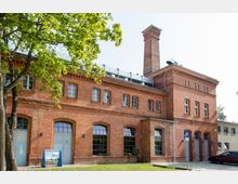 Backsteingebäude im industriellen Stil mit hohen Fenstern und einem markanten Schornstein, umgeben von Bäumen und einem gepflasterten Vorplatz. Im Vordergrund ist ein kleiner Parkplatz mit einem Auto und einem Parkplatzschild zu sehen.