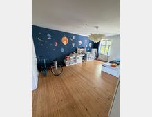 Großes Kinderzimmer mit hellem Holzboden und dunkelblauer Akzentwand, die mit Planeten- und Sternmotiven gestaltet ist. Entlang der Wand stehen weiße Regale und ein Schreibtisch; neben einem großen Fenster mit Heizkörper stehen ein Bett sowie abgestellt ein Fahrrad und ein Roller.