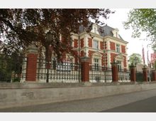 Historische Villa mit rotem und weißem Fassadenmuster, steilem Schieferdach und dekorativen Fenstereinfassungen, umzäunt von einem schmiedeeisernen Zaun auf gemauerten Säulen; im Vordergrund befindet sich ein Gehweg aus Kopfsteinpflaster.