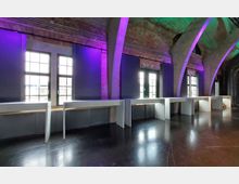 Industrieartig gestalteter Innenraum mit Backsteinwänden, großen Fensterfronten und markanten Betonbögen. Weiße, minimalistische Tische stehen vor den Fenstern, und farbige Beleuchtung in Grün und Violett akzentuiert die Decke und Wände.