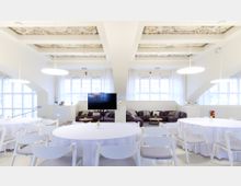 Heller, modern gestalteter Innenraum mit hohen Fenstern, weißer Einrichtung und runden Tischen mit weißen Tischdecken. Im Hintergrund eine gemütliche Sitzecke mit dunklen Sofas, kleinen Holztischen und verzierten Deckenpanelen.