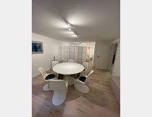 Heller Innenraum mit einem runden weißen Tisch und sechs weißen Stühlen mit schwarzen Sitzpolstern auf einem hellen Holzboden. Im Hintergrund sind ein Raumteiler, geschlossene Türen und eine schlichte Wanddekoration zu sehen.