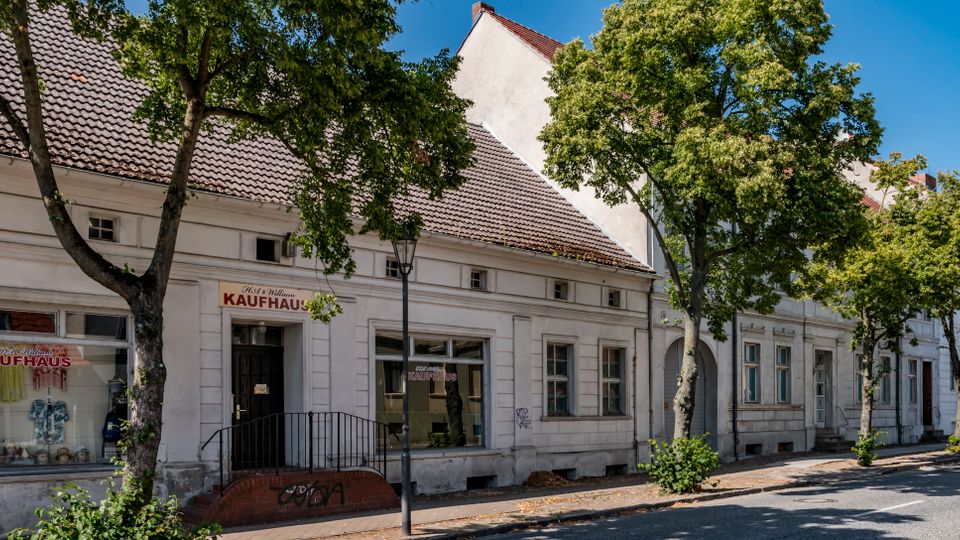 Straßenansicht einer Reihe niedriger, weiß verputzter Altbauten mit Ziegeldächern und großen Schaufenstern, darunter ein Geschäft mit dem Schild „Kaufhaus“. Entlang des Gehwegs stehen mehrere Bäume und eine Straßenlaterne, die Straße verläuft ruhig vor den Gebäuden.
