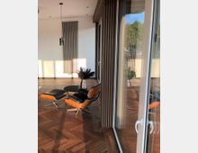 Moderner, minimalistischer Wohnraum mit Holz-Fischgrätenboden, einer bodentiefen Glasfront, einer Stehlampe, einem Design-Sessel mit passendem Hocker aus Holz und schwarzem Leder sowie einer Vase mit Trockenblumen. Im Hintergrund eine weiße Wand mit vertikalem Heizkörper.