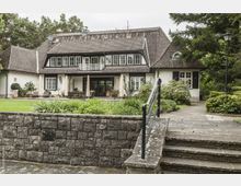 Großes, zweistöckiges Landhaus mit weißer Fassade, dunklem Ziegeldach und zahlreichen Fenstern, umgeben von gepflegtem Garten und Bäumen. Eine Terrasse mit Gartenmöbeln sowie ein gepflasterter Weg und eine Steinmauer führen zum Haupteingang.