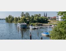 Hafenbereich an einem See mit Anlegestellen für Boote, einem schwimmenden Gebäude und mehreren Motorbooten. Im Hintergrund sind dichte Baumreihen, moderne Gebäude und eine Kirche mit zwei Türmen zu sehen.
