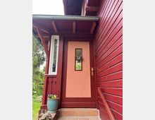 Rote Holztür eines Hauses mit einem kleinen rechteckigen Fenster, geschmückt mit einem Kranz. Die Fassade des Hauses besteht aus rot gestrichenen Holzlatten, daneben steht ein Blumentopf mit bunten Blumen auf einer gefliesten Stufe.