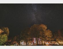 Nachthimmel mit deutlich sichtbaren Sternen und der Milchstraße. Im Vordergrund befinden sich beleuchtete Bäume und Gebäude, darunter ein erleuchtetes Auto in einer parkähnlichen Umgebung.