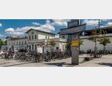 Bahnhofsvorplatz in Bernau bei Berlin mit zahlreichen geparkten Fahrrädern, einem historischen Bahnhofsgebäude im Hintergrund und einer modernen Stele mit Temperaturanzeige und Beschilderung. Der Platz ist gepflastert und von Bäumen gesäumt, mit Personen in der Umgebung und einem klaren Himmel darüber.