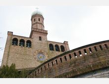 Turm einer Backsteinkirche mit Rundbogenfenstern, einer Uhr mit Rosenmotiv und einem Kuppeldach mit Kreuz an der Spitze. Im Vordergrund ist ein gemauerter Rundbogenzaun zu sehen.