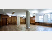 Großer leerer Raum mit Holzboden im Fischgrätenmuster, hellen Wänden und mehreren rechteckigen Fenstern auf der rechten Seite. Die Wände sind teilweise mit Holztäfelungen verkleidet, und es gibt zwei freistehende weiße Säulen sowie hängende Leuchtstofflampen an der Decke.