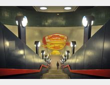 U-Bahn-Unterführung mit blauer Wandverkleidung und roten Handläufen, beleuchtet durch entlang der Treppe angebrachte runde Lampen. Die Decke ist mit einem großen gelb-roten Wandgemälde verziert, das ein Cartoon-Gesicht zeigt.