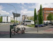 Straßenecke mit Schildern „Bayerischer Platz“ und „Meraner Straße“, umgeben von mehrstöckigen Wohn- und Geschäftsgebäuden. Im Vordergrund stehen zwei Fahrräder an einem Mast, während Autos, ein kleines Parkstück und eine U-Bahn-Eingangsstation im Hintergrund sichtbar sind.