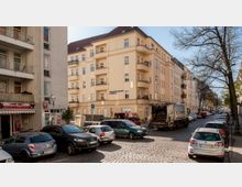 Straßenszene in einer Stadt mit gepflasterter Straße, parkenden Autos und einem mehrstöckigen Wohnhaus mit hellgelber Fassade und Balkonen im Hintergrund. Rechts und links der Straße stehen Bäume, die teilweise blättrig sind, während im Vordergrund ein Müllfahrzeug zu sehen ist.