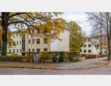 Mehrfamilienhäuser mit cremefarbenen Fassaden in einer von Bäumen und Hecken umgebenen Wohngegend. Im Vordergrund sind ein gepflasterter Gehweg, eine Bank und ein Straßenschild mit der Aufschrift "Jungfernstieg" zu sehen.