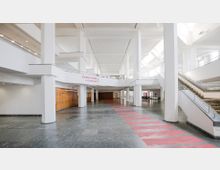 Großzügiger, heller Innenbereich eines modernen Gebäudes mit hohen weißen Säulen, einer offenen Galerie und großen Fenstern, die Tageslicht hereinlassen. Im Vordergrund befinden sich ein mit roten Streifen markierter Boden und an der Seite ein Bereich mit Schließfächern; rechts führen breite Treppen mit Geländern nach oben.