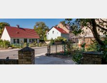Blick auf eine ruhige Wohnstraße mit kleinen Einfamilienhäusern, darunter ein weißes Haus mit rotem Ziegeldach und grünen Fensterläden. Im Vordergrund ein Garten mit grünem Metallzaun, umgeben von Bäumen und Blumenbeeten.