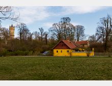 Eine gelbe, einstöckige Hütte mit rotem Ziegeldach steht in einem Park mit einer offenen Wiese im Vordergrund, umgeben von Bäumen. Im Hintergrund ist ein historischer Turm auf einem bewaldeten Hügel zu sehen.