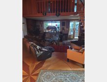 Wohnzimmer mit Holzboden und Teppich, möbliert mit Sofas und Sesseln. Im Hintergrund große Fensterfront mit Blick auf eine dekorierte Terrasse und hölzerne Treppe, die zu einer Galerie im oberen Bereich führt.