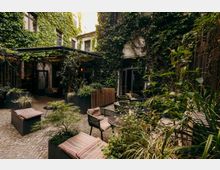 Innenhof-Terrasse eines Gebäudes mit kopfsteingepflastertem Boden, kleinen runden Tischen und Stühlen sowie Sitzbänken mit gestreiften Polstern. Die umgebenden Fassaden sind dicht mit Efeu bewachsen, ergänzt durch viele Pflanzkübel und eine überdachte Pergola am Eingang.