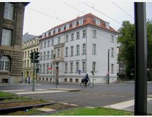 Ein mehrstöckiges, weißes Gebäude mit rotem Schrägdach an einer Straßenkreuzung, umgeben von weiteren Stadthäusern. Im Vordergrund sind Straßenbahnschienen, Gehwege, eine Fahrradfahrerin und eine Ampel zu sehen, im Hintergrund steht ein Baum.