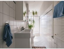 Helles Badezimmer mit weißen Fliesen an Wänden und Boden, einem Waschbecken mit Spiegel, einer bodentiefen Dusche im Hintergrund und einem Heizkörper mit Handtuch daneben. Pflanzen und ein bunter Teppich sorgen für dekorative Akzente.