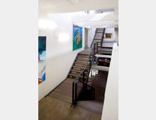 Offener Treppenbereich in einem modernen, weißen Gebäude, mit Holzböden und Geländern aus Metall. Die Wände sind mit großformatigen, bunten Gemälden dekoriert, und im Hintergrund ist ein Bürobereich mit Regalen und Schreibtischen zu sehen.