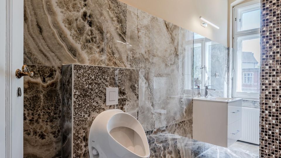 Modernes Badezimmer mit großformatigen, marmorierten Steinfliesen an Wänden und Boden, Urinal und wandhängendem Waschtisch. Rechts sorgt ein großes Fenster für Tageslicht; eine Mosaikfliesenwand und eine Leuchte sind sichtbar.